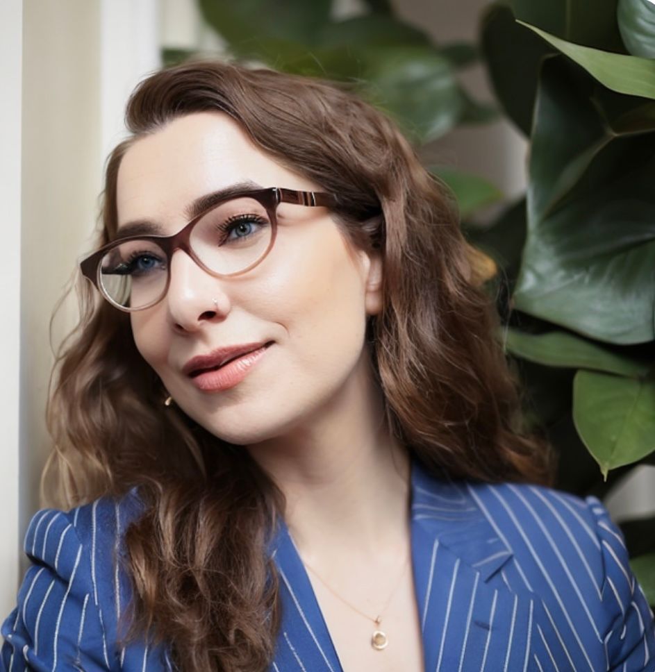 Portrait of Kara Hokes, PhD, a clinical psychologist with wavy brown hair and tortoiseshell glasses, smiling in front of a leafy green plant